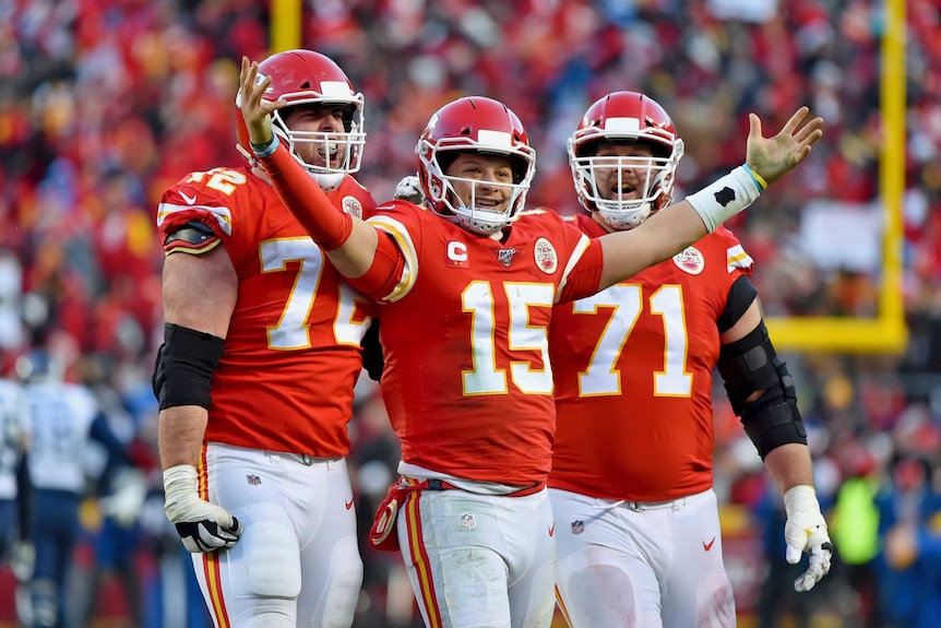Surrounded by two teammates, Patrick Mahomes holds both arms up and smiles.