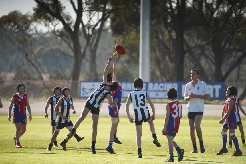 Children wearing black and white and red and blue jerseys gather around two players reaching for a ball on the footy oval
