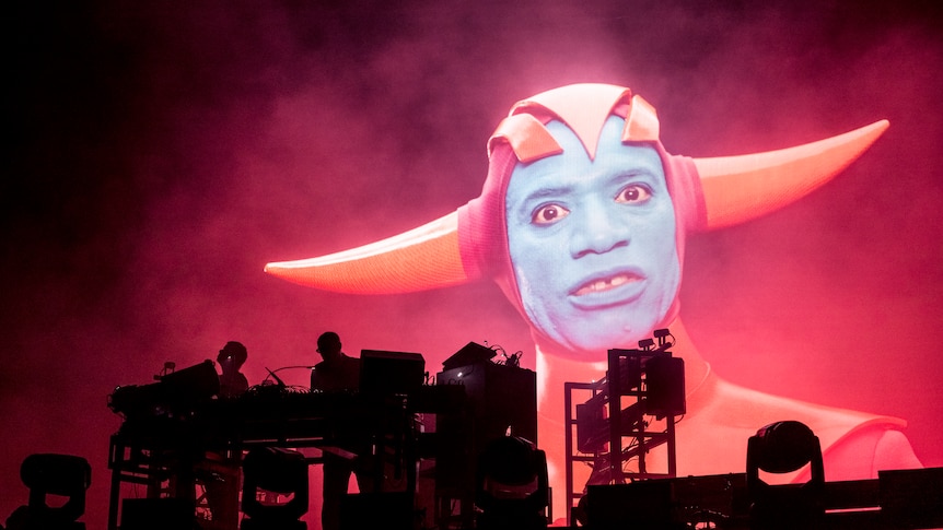 A creature with a blue painted face and red horns on a screen behind UK duo the chemical brothers