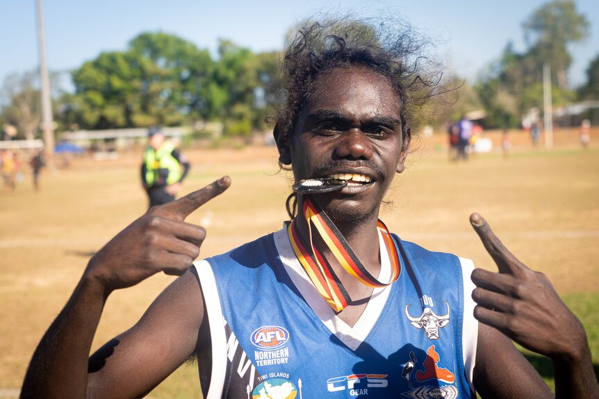 A young Indigenous footy player holds his victory medal in his mouth and points at it smiling