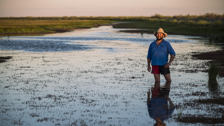 A man stands knee deep in water, standing among wetlands at sunset with a smile on his face.
