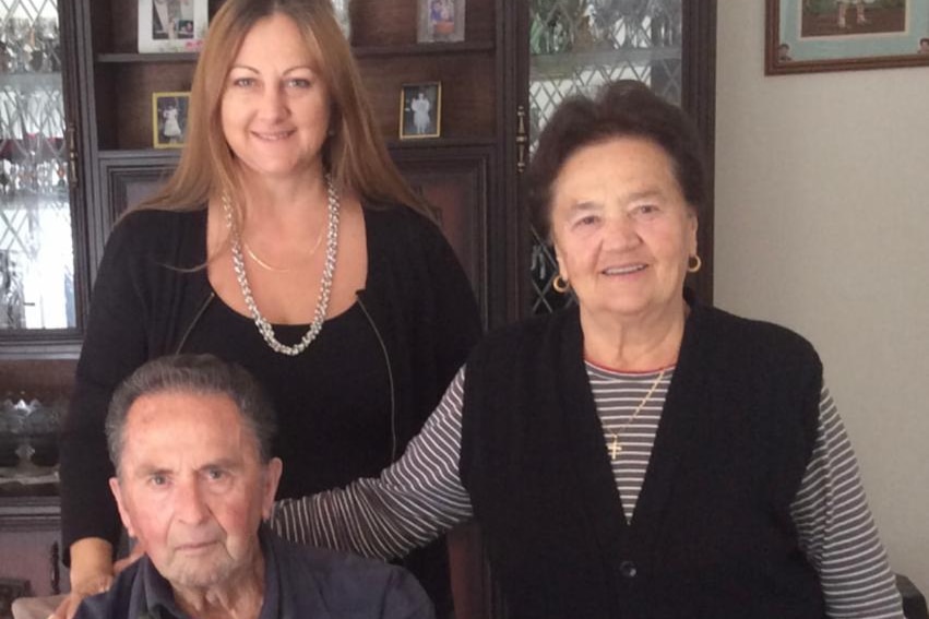 Two women stand, one has arm around man who is seated in chair with arms folded.