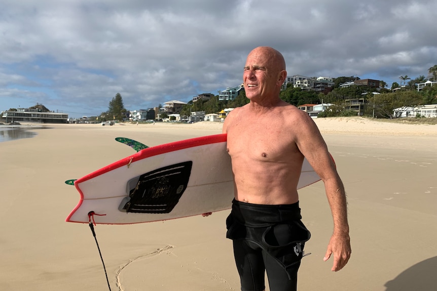  A man with his wetsuit rolled down carries a surfboard on a beach, semi-cloudy sky, houses, trees in backgound.