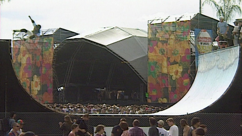 A video still as a skateboarder ascends a half pipe with a large music stage in background.