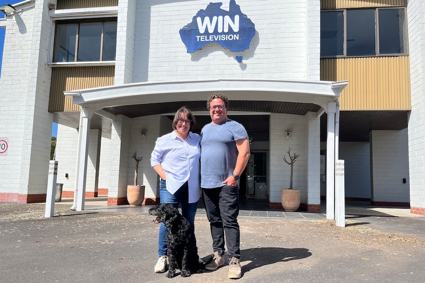 A woman, a man and a dog standing outside a building with a logo for WIN TV on it.