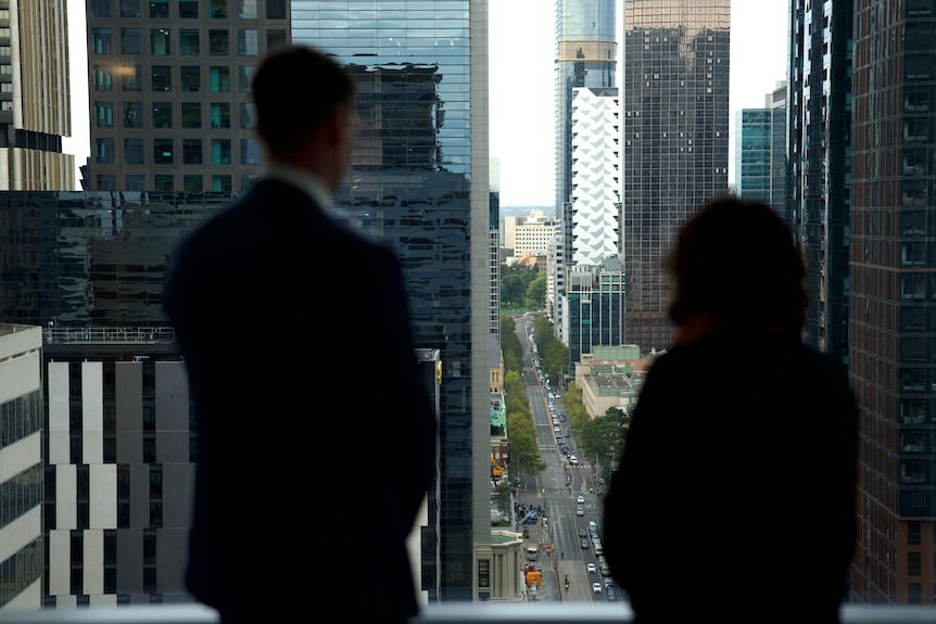 Joel Phibbs and Kimi Nishimura stand by a large high-rise window with the traffic visible on the street below