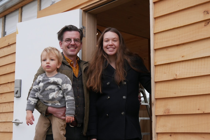 Family of three, a man holding a young boy and the mother next to them in the doorway of a new home.