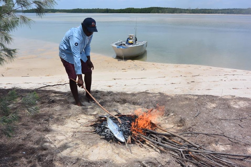 A young Indigenous man cooking fish on an open fire at a beach.