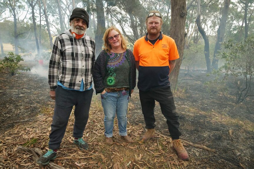 A women stands between two men in a stand of trees with smoking ground behind them.