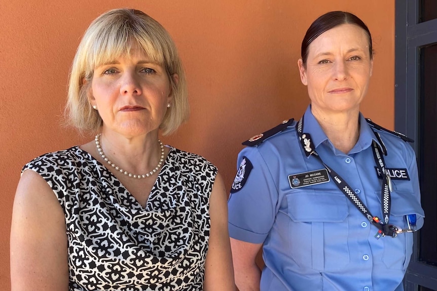 Two women stand against a wall, one wearing civilian clothes and one a police uniform.