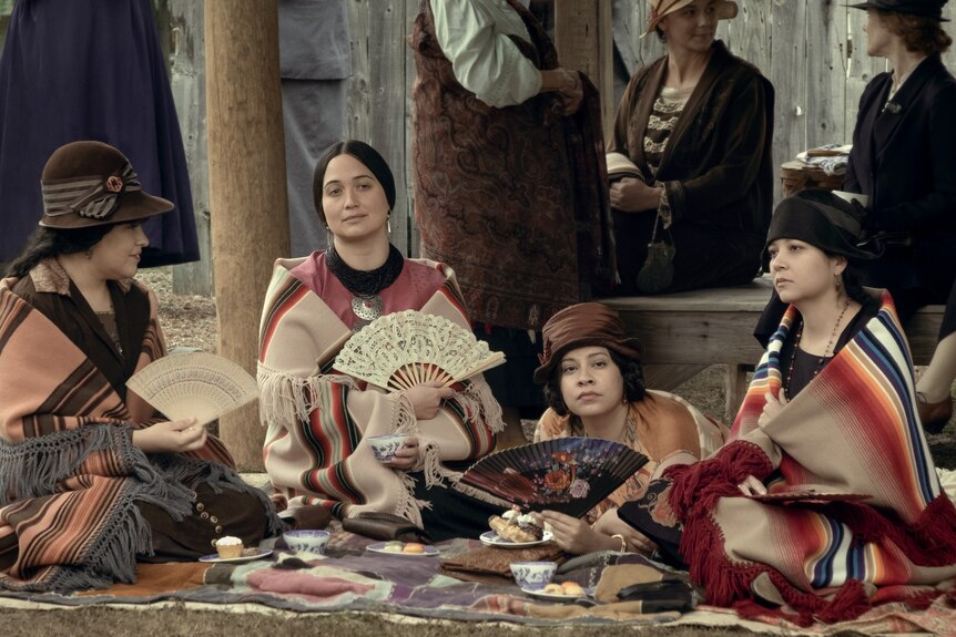A film still showing four Native American women sitting on the ground, wearing traditional shawls and some holding fans