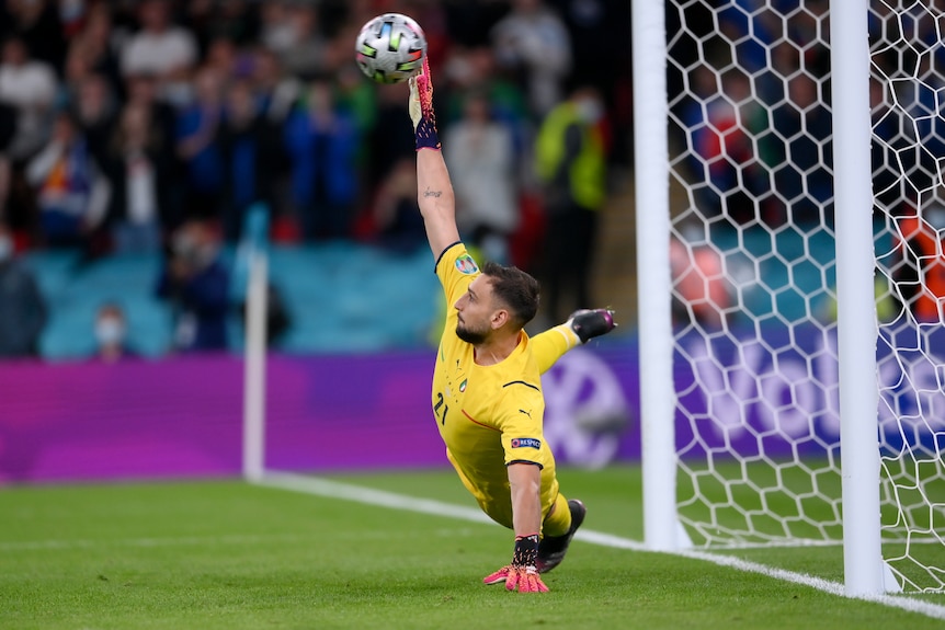 A goalkeeper has one hand on the turf and the other stretched above him to save a penalty as he dives sidewards.