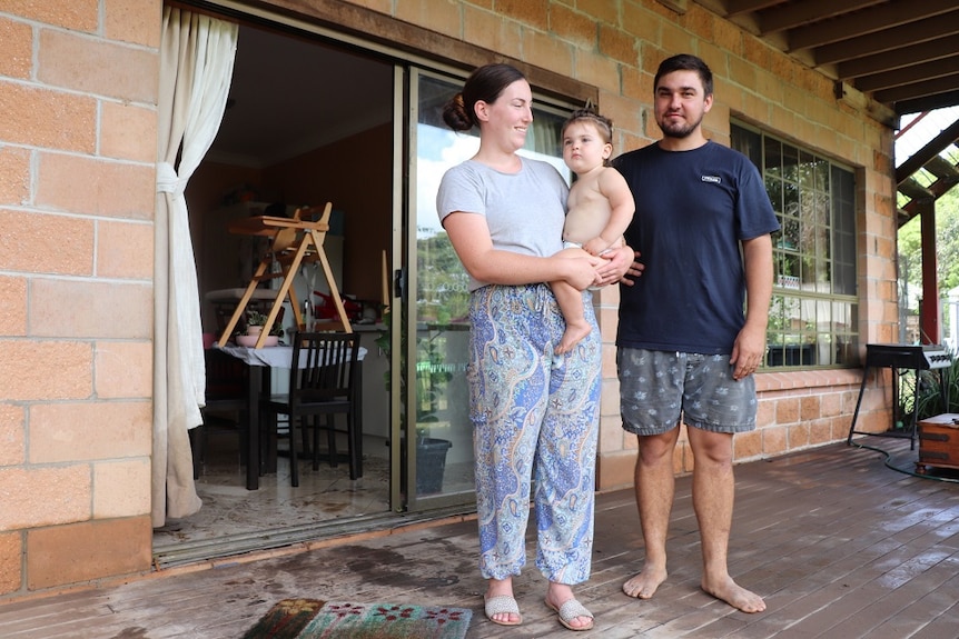 Emily Campbell with fiance Jesse and baby Eden outside their home in Nana Glen to day after it was inundated by floodwater.
