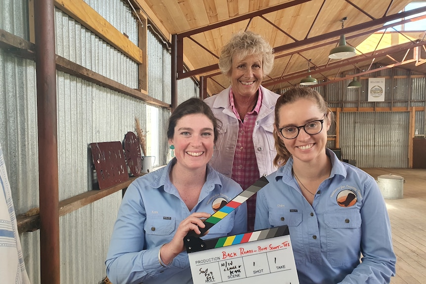 Two women with dark hair sit holding a clapboard. A woman with grey hair stands behind. They're in a shed.