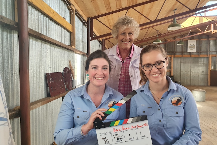 Two women with dark hair sit holding a clap board, a woman with grey hair stands behind. They're in a shed. 