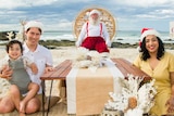 A man and woman sit with their small child at a low table on the beach, with Santa in a chair at the end.