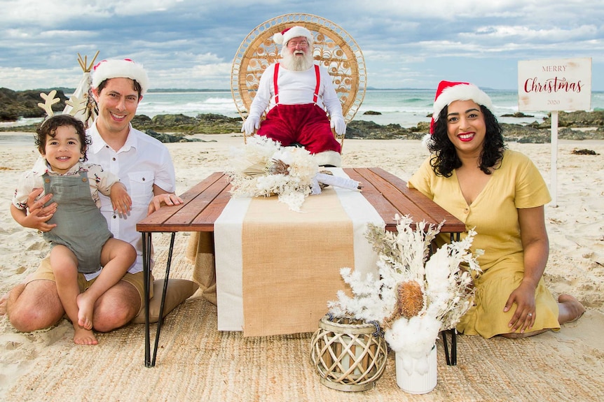 A man and woman sit with their small child at a low table on the beach, with Santa in a chair at the end.