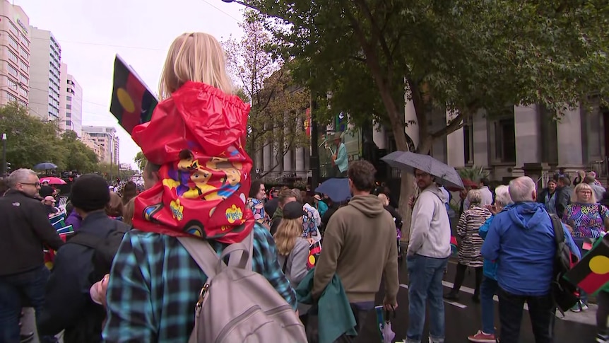 A small child sitting on the shoulders of an adult waves an Aboriginal flag, she's in a crowd of people