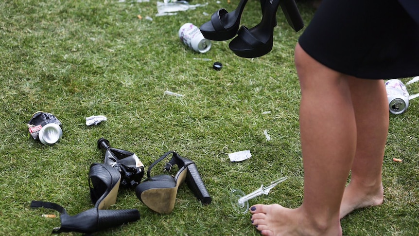 Woman stands barefoot, heels in hand amongst shoes and litter on the grass