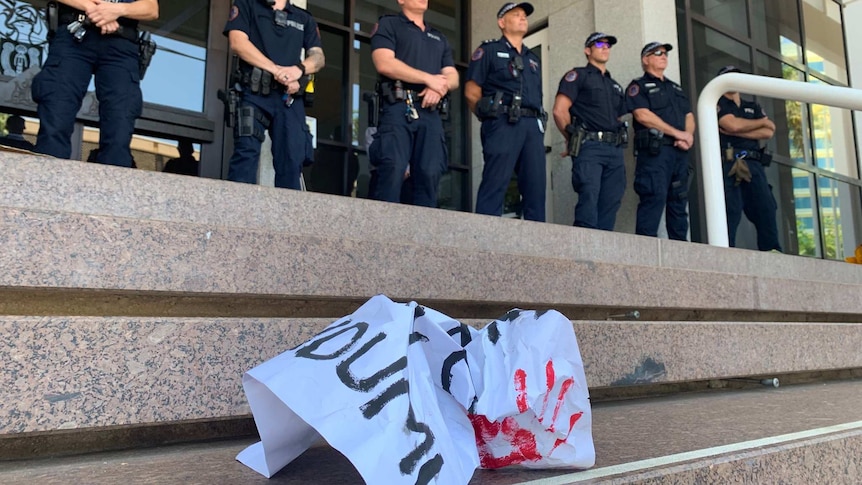 A line of police officers stand on at the top of some wide stone steps with a crumpled poster in the foreground.