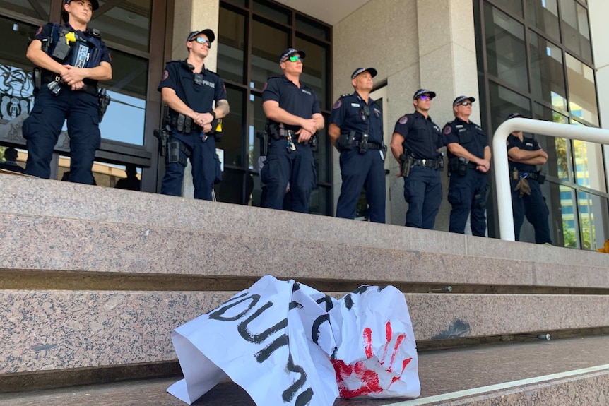 A line of police officers stand on at the top of some wide stone steps with a crumpled poster in the foreground.