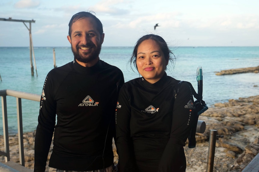 Narius and Hope in black wetsuits, smiling, standing side by side, bird, gantry and ocean behind.