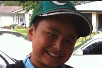 A young boy stands in a pale school uniform and cap, smiling. 