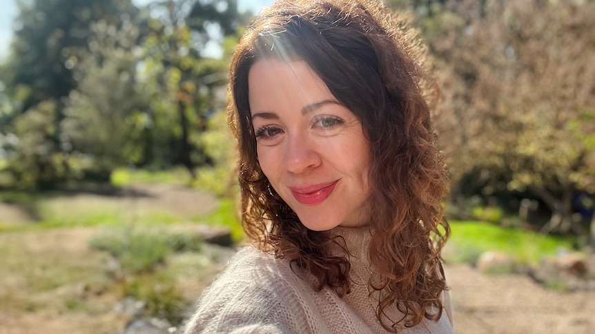 Head and shoulders image of a woman smiling in the sunshine in the outdoors.