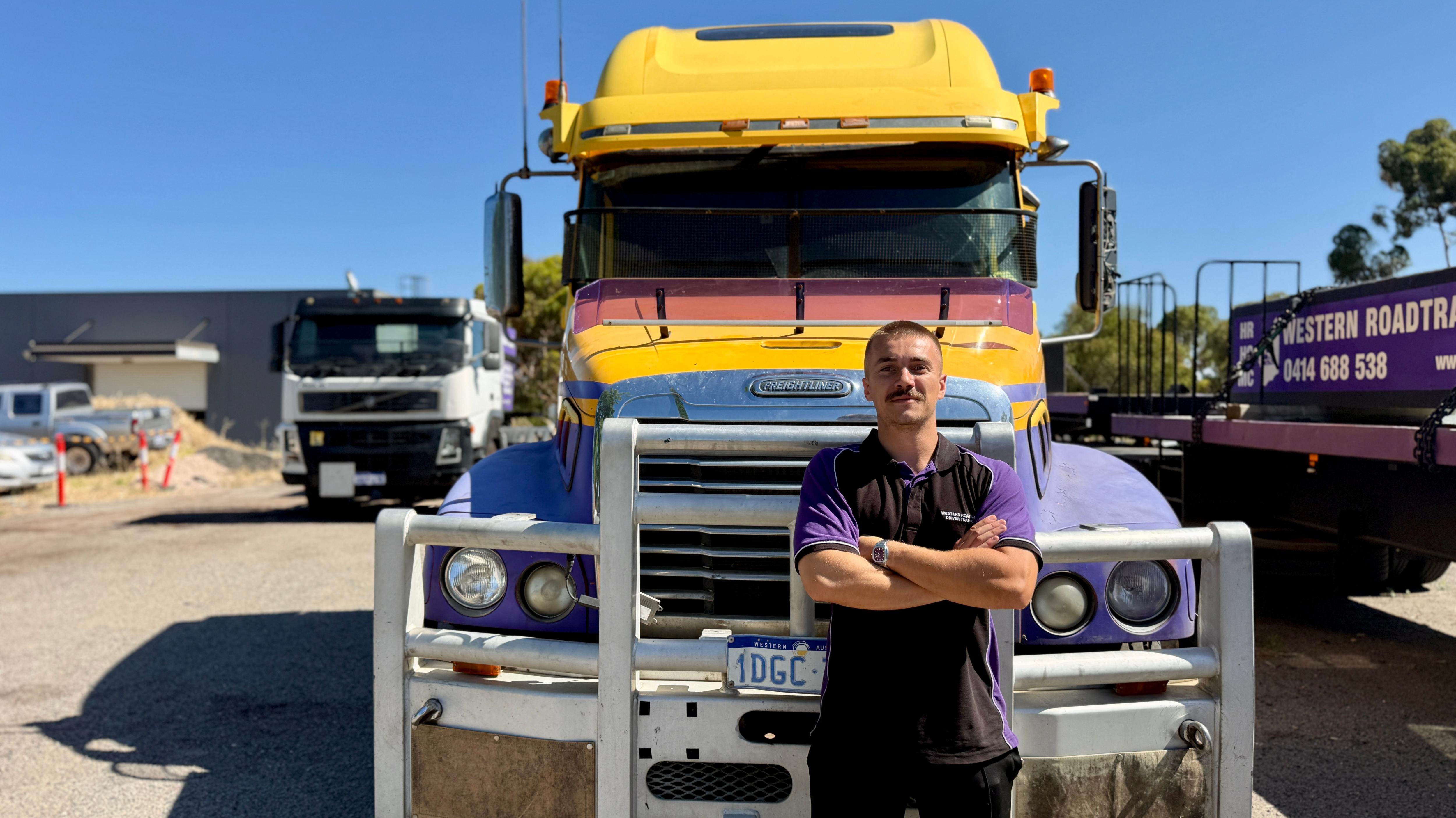 Un joven se para frente a un camión grande con los brazos cruzados.