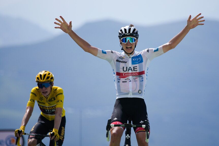 A rider in a white jersey smiles and raises his arms in triumph as he finishes a stage ahead of a rider in the yellow jersey.