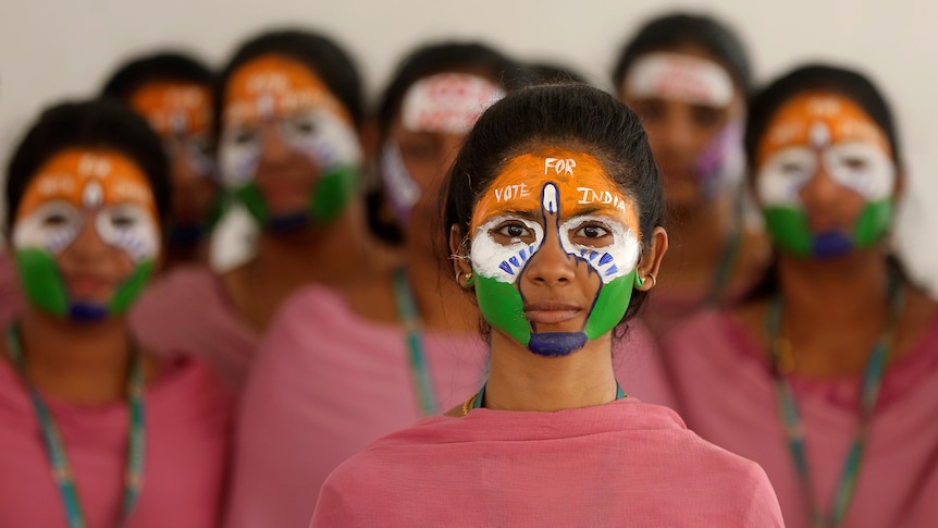 Young with the colours of the Indian national flag painted on their faces wear red tops and stare at the camera as they pose.