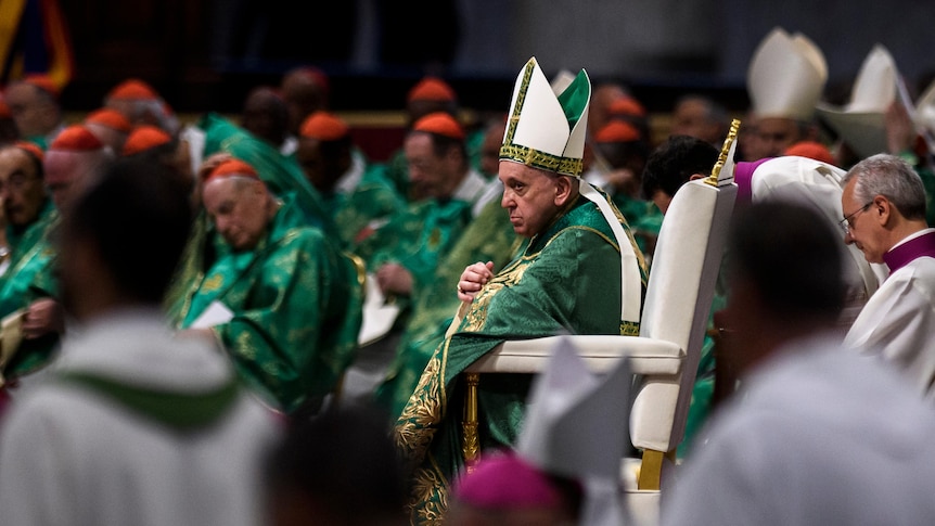 side view of pope francis in green robes with gold pattern. cardinals wearing similar colour robes in background