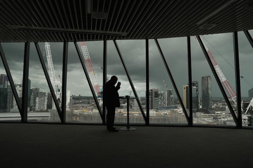 A silhouetted man stands at a series of windows looking out over the city. He cleans his glasses.