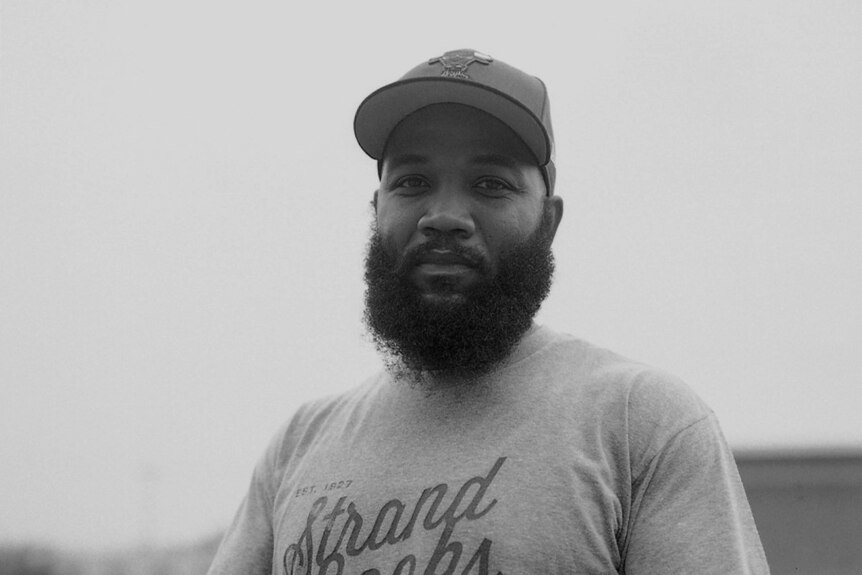 Dr A.D. Carson, a black and white photo of a black man with beard wearing a baseball cap and t-shirt