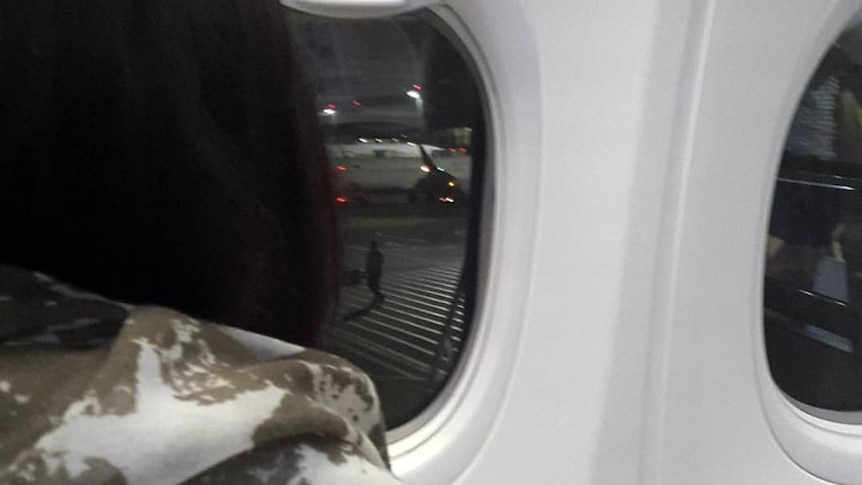 Woman with black hair (assumed to be Corby) looks out of an airplane window to the tarmac outside.