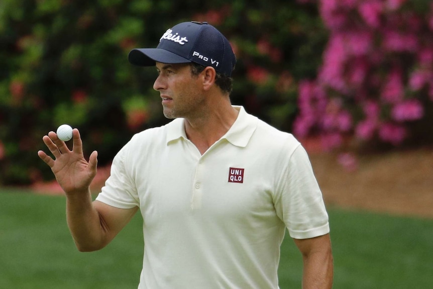 Adam Scott stands holding a golf ball looking off to his right, wearing an off-white polo shirt and dark blue cap