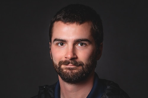 A young man with short dark hair and a beard  looks at the camera.