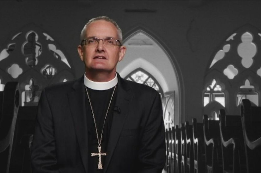 A man in black religious robes is standing behind black church pews.