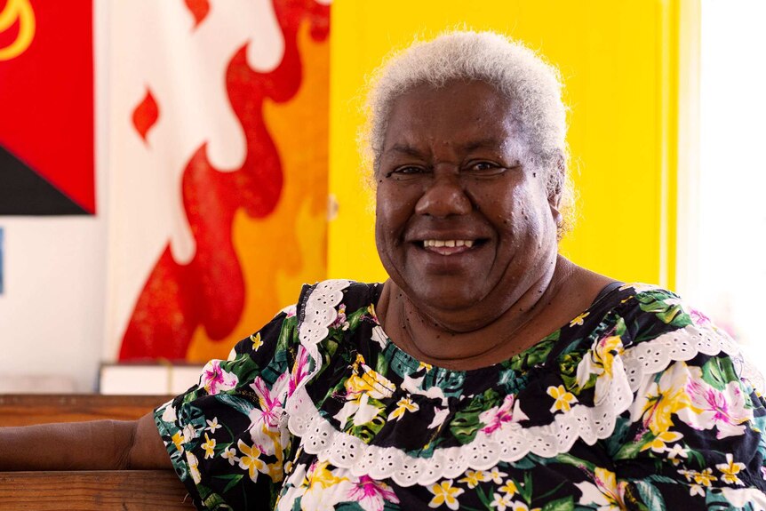 Midshot of woman with white hair smiling to camera against colourful yellow and red background.