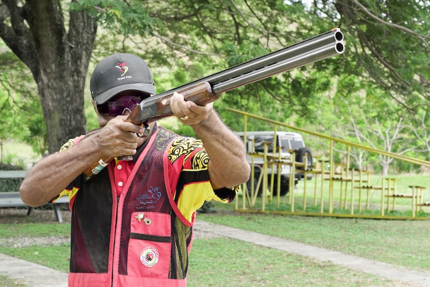 A Black man wearing sunglasses and a black cap holds a gun pointed in the sky with trees in the background.