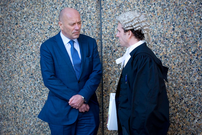 Two white men stand outside a courtroom. One is bald and wearing a blue suit, the other is a barrister wearing a robe