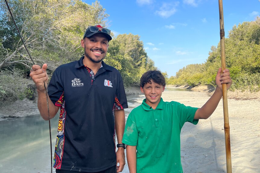 A man and a boy both holding long sticks while smiling at the camera and standing in a sandy creek.