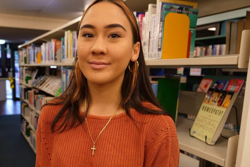 A girl with olive skin and dark hair stands smiling in a library aisle wearing an orange jumper and gold cross necklace.