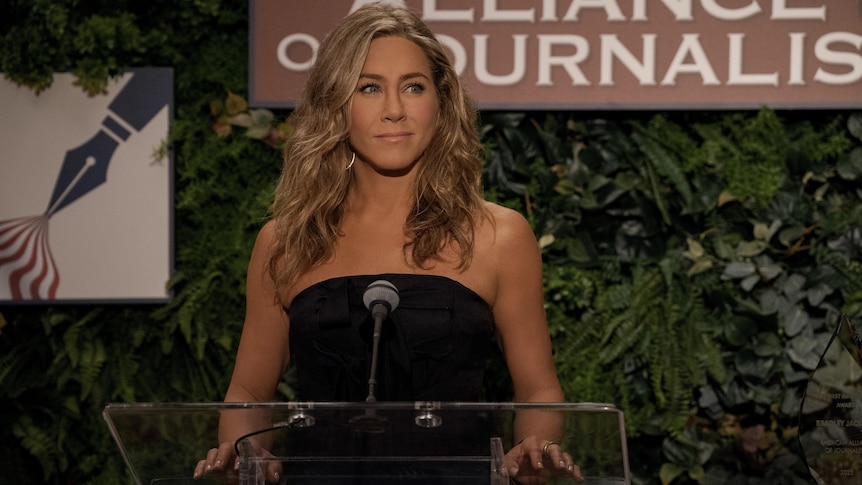 A white woman with brown wavy hair is standing at a podium, she looks calm and collected