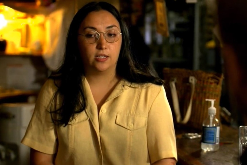 A woman in a light blouse and glasses sits in front of an empty bar.