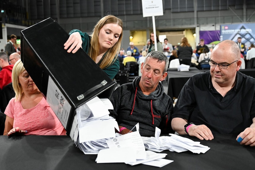 Three people sitting at a table, while a woman tips a large box of paper votes out in front of them