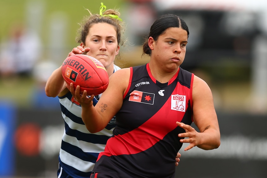 An Essendon AFLW player holds the ball in her right hand in front of a Geelong opponent.
