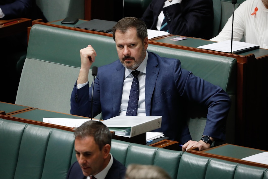 A dark-beared Ed Husic is sitting in parliament, holding a pen, his left arm braced against his desk.