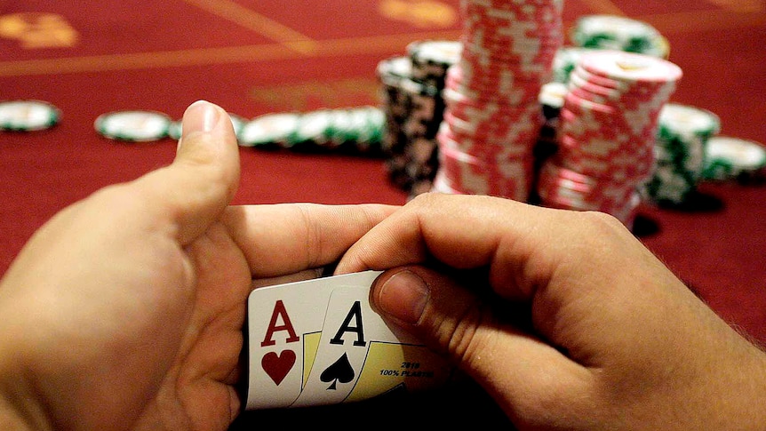 Two ace playing cards are held up in front of piles of poker chips stacked on a gaming table.
