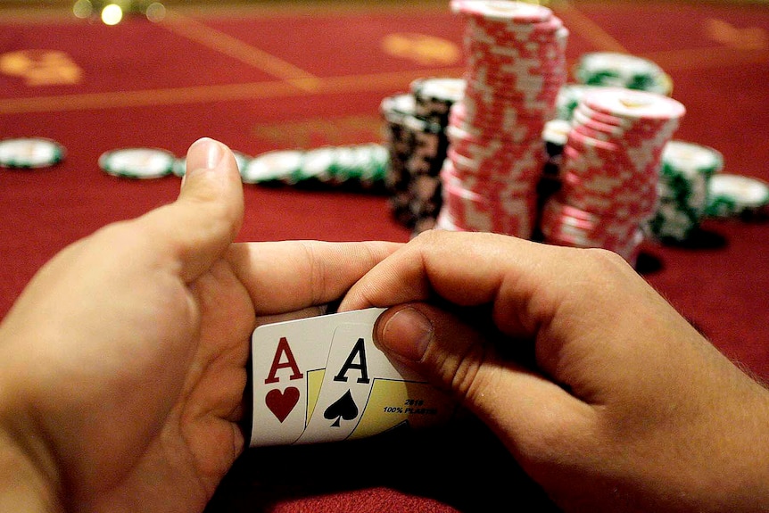 Two ace playing cards are held up in front of piles of poker chips stacked on a gaming table.
